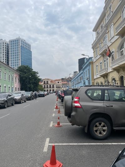 The image captures a street scene in Luanda, Angola, under an overcast sky during the daytime. The perspective is down a wide asphalt road marked with white parking lines. A row of parked cars, predominantly SUVs, lines the right side of the street, delineated by orange traffic cones. The closest vehicle is a gray SUV with a spare tire mounted on its rear.
On the left, a varied architectural landscape unfolds. A prominent multi-story building, painted light yellow with ornate white window frames and balconies, suggests colonial-era design. From this building, a distinctive Angolan flag, red and black with a yellow emblem (machete, cogwheel, star), is visibly hanging. Further along the street, other buildings, including a light blue structure and several in pastel shades, line the road. In the background, modern high-rise buildings rise above the older structures, illustrating the urban development of the city. A small cluster of green trees is also visible. The sky is uniformly gray, indicating a cloudy day. No people or legible text are visible within the frame.