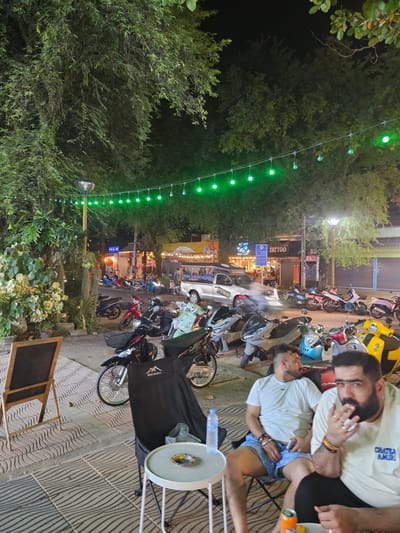 This image depicts a nighttime scene on a street in Ban Klong Haeng, Thailand. The street is lined with various parked motorcycles, suggesting a busy area or perhaps a gathering spot. Overhead, a string of green lights illuminates the scene, casting a festive glow. In the foreground, two men are seated, seemingly engaged in conversation or relaxation. One man, wearing a light-colored t-shirt and blue shorts, is looking towards the camera with a relaxed expression. The other man, with a beard and wearing a similar t-shirt, is touching his face, with a ring on his finger. Behind them, a black tarp is draped over something, possibly a chair or equipment. A small white table with a circular top sits nearby, with a bottle of water and a can on it. A wooden easel with a blackboard stands on the sidewalk.

Further down the street, a tattoo parlor is visible with illuminated signage. Other shops and businesses are also present, indicated by their lights and awnings. The presence of numerous motorcycles points to a location where transportation is common and perhaps a popular social hub. The overall atmosphere appears casual and lively, typical of a warm evening in a Southeast Asian locale. The time of day is clearly night, and the weather appears to be clear.
