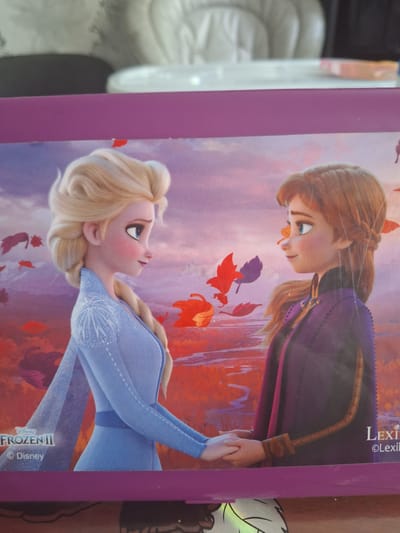 The image is a close-up of a purple box, likely a lunchbox or storage container, featuring characters from Disney's "Frozen II." The main subjects are Elsa and Anna, the two protagonists, depicted in profile facing each other. Elsa, on the left, has blonde hair styled in a braid and wears a light blue dress with intricate white patterns. Anna, on the right, has auburn hair and wears a dark purple cloak over a lighter purple garment. Both characters are holding hands. The background of the image is an autumnal landscape with a purple and orange sky, stylized trees, and falling leaves. The "Disney Frozen II" logo is visible in the bottom left corner of the image, along with the "Disney" copyright mark. A "Lexi" logo is also present in the bottom right corner. The box appears to be resting on a surface, with the background out of focus, suggesting an indoor setting. There is no visible indication or context that would place this image in Bochum, Germany.