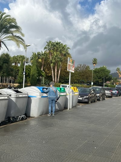 A daytime street scene in Pineda de Mar, Spain, depicts a female individual, seen from behind, with gray hair, wearing a denim jacket and blue jeans, actively placing an item into a blue-lidded municipal recycling bin. Five large, grey recycling bins are arranged in a row along a dark grey paved sidewalk, featuring lids of varying colors: two grey, one blue, one green, and one yellow. To the right, a line of at least six cars is parked parallel to the curb on an asphalt road; the closest vehicle is a dark grey Volkswagen Polo. A tall grey street lamp is positioned behind the bins to the left, and a white pole with a multi-colored information sign stands between the green and yellow bins. The background is lush with dense green vegetation, including numerous prominent palm trees, suggesting a park or landscaped area. The sky above is partly cloudy with patches of blue visible. Small white debris is scattered on the sidewalk.