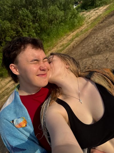 A close-up selfie shows two young people outdoors, with a sandy incline and green foliage in the background. The person on the left, with short dark hair and closed eyes, is being kissed on the cheek by the person on the right. The person on the left wears a red shirt and a blue towel with an embroidered bird. The person on the right wears a black tank top and a necklace with a cross. The lighting suggests it is late afternoon. The location is likely a natural area near Рябинино, Russia.