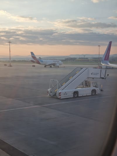 The image shows a view from an aircraft window at Stuttgart Airport (STR) in Echterdingen, Germany. Two airplanes are visible on the tarmac: a Eurowings Airbus A320 and a Wizz Air airplane. A passenger boarding bridge with "Stuttgart Airport" branding is positioned on the tarmac near the window. The sky has a mix of blue and pink hues, suggesting either sunrise or sunset. Ground equipment and infrastructure are visible in the background, including lighting poles and other airport vehicles. The ground is concrete with painted lines.