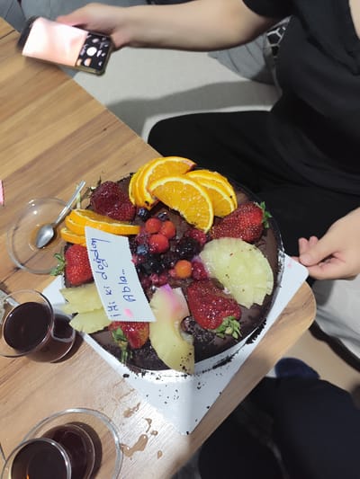 A chocolate cake topped with strawberries, blueberries, raspberries, pineapple, and orange slices is placed on a wooden table. A white card with blue handwritten text that reads "İyi ki doğdun Abla..." is inserted into the cake. A spoon rests in a small glass bowl to the left of the cake, and two glasses filled with dark liquid are visible in the foreground. A person's hand is holding a smartphone, with the screen illuminated and showing colorful images, in the upper left corner of the frame. The background shows a person wearing a black shirt. The image appears to be taken indoors, possibly in a residential setting, and given the text on the card, it is likely a birthday celebration. The city of Kâğıthane, Turkey, is mentioned in the context of this scene.
