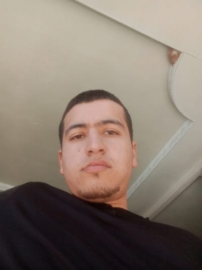 A young adult male is depicted indoors, within a building located in Kasan, Uzbekistan. The subject, with short dark hair, dark prominent eyebrows forming a unibrow, and light facial hair on the chin and upper lip, occupies the lower portion of the frame. He wears a dark, unidentifiable upper garment. The camera perspective is low-angle, looking up at the subject, who maintains a direct, neutral gaze. The background reveals a light-toned ceiling with architectural molding in the upper right, exhibiting a curved ornamental design. A small, irregular cluster of material, consistent with an insect nest or debris, is present in the corner where ceiling planes and molding converge. No specific actions or interactions are observed.