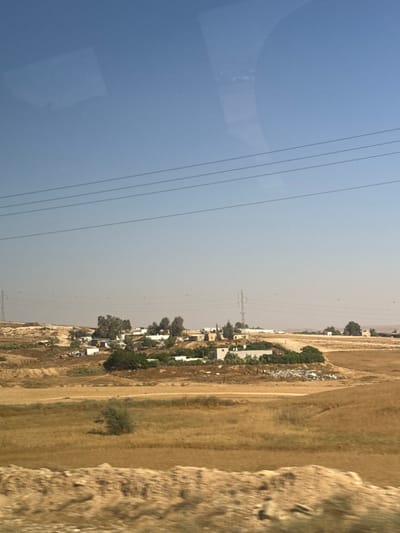 This media file shows a view from a vehicle, possibly a train, looking out at a rural landscape. The scene is divided by power lines that stretch across the frame. On the left side, the sky is a clear, pale blue, reflecting the time of day as likely daytime, perhaps late morning or afternoon. There are some indistinct, blurry shapes on the left side of the sky which might be reflections on the glass of the vehicle.
The right side of the image depicts a dusty, dry landscape with patches of vegetation and a small settlement. The settlement consists of several low-rise buildings with white or light-colored roofs, scattered among trees. A dirt road winds through the area. In the distance, there are power poles and transmission lines, indicating the presence of electrical infrastructure. The ground is covered in dry grass and sparse foliage, suggesting a warm, arid climate. The overall impression is of a somewhat underdeveloped or agricultural area.
There are no visible people in the frame, nor any specific objects that stand out beyond the general elements of the landscape and infrastructure. The activity is static; the image captures a view of the passing scenery. The lighting is bright, consistent with daylight. There are no explicit cues to a specific time of day beyond general daytime, and no weather phenomena are apparent. The location context provided suggests this is in Israel, within the al-Qasoum Regional Council, which aligns with the dry, rural appearance. No text is visible in the image.
