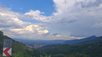 This is a high-angle, wide-shot photograph taken from a hillside overlooking a valley with a town. The sky dominates the upper half of the image, filled with a mix of bright white and darker, stormier grey clouds. The sun is likely behind the clouds, casting a diffused light on the landscape.

In the foreground, on the left side of the image, a roadside sign is visible. It is a rectangular grey sign mounted on a metal pole, with a red and white chevron sign at the bottom, indicating a sharp turn to the left. Lush green trees and foliage cover the steep hillside in the immediate foreground and frame the lower edges of the scene.

The middle ground reveals a sprawling town nestled within the valley. The buildings appear to be a mix of residential and commercial structures, with a denser concentration in the center. The town is surrounded by rolling green hills.

In the background, a series of mountain ranges recede into the distance, fading in color from dark green to a hazy blue. The overall impression is one of a scenic, natural landscape with a human settlement integrated into it. The location is identified as Dobrotino, Bulgaria.