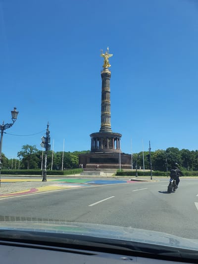 A wide-angle shot captures the Victory Column in Berlin, Germany, on a clear, sunny day. The iconic monument, topped with a golden statue of Victoria, stands tall against a bright blue sky. The column's detailed structure, with its ornate carvings and tiered design, is clearly visible.
In the foreground, a person rides a motorcycle on a street painted with colorful stripes, resembling a rainbow. The motorcycle has a rider wearing a helmet and dark clothing. The street is paved with asphalt, and white lines mark the traffic lanes.
Beyond the monument, lush green trees form a dense line, suggesting a park or well-maintained urban landscape. Lampposts and traffic signals are also visible along the street. The scene appears to be during daylight hours, with the sun casting distinct shadows. The overall impression is one of a vibrant and active urban environment in Berlin.
