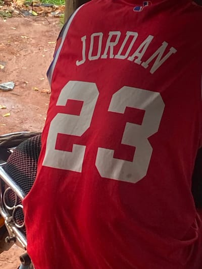 A red sports jersey, emblazoned with the name "JORDAN" printed vertically in white capital letters and the large white number "23" horizontally, dominates the frame. The jersey features a subtle blue and white trim along its visible edge, and a small, partially obscured blue and red emblem or logo can be seen on the upper left side, near the 'J' in Jordan.
The jersey appears to be draped over or resting on an object, possibly a vehicle such as a motorcycle or scooter, indicated by metallic components and black mesh material visible in the bottom right corner of the image. Below the jersey and vehicle, the ground is an earthy brown, suggesting an unpaved surface, scattered with dried leaves and small rocks.
The setting is outdoors, likely a casual street or yard area, characteristic of an everyday environment in Auchi, Nigeria. The lighting indicates it is daytime. No individuals are visible, and no specific activity is occurring. The image is presented with a rotated perspective.