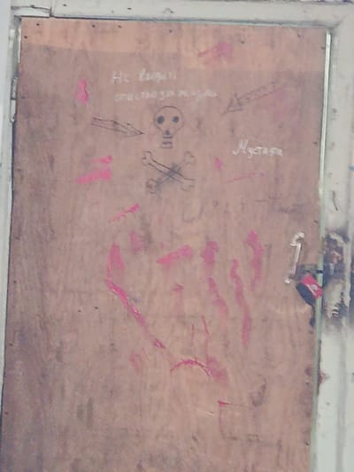 The image shows a wooden door with various drawings and markings in a building, likely located in Sokuluk, Kyrgyzstan. In the upper left portion of the door, white text reads "Не входить" which translates to "Do not enter". Below this text, a crudely drawn skull and crossbones is depicted. To the right of the skull and crossbones, in white, is the word "Мустафа". There are also several pinkish-red smudges and handprint-like markings scattered across the door. A black lock is attached to the right side of the door. The overall condition of the door suggests it is in a neglected or abandoned space.