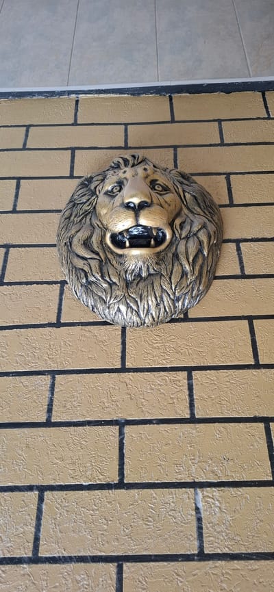 The image shows a close-up, vertical shot of a decorative lion's head mounted on a brick-patterned wall. The lion's head is rendered in a metallic gold and bronze finish, with its mouth open in a roaring or snarling expression. Its mane is sculpted with dramatic, flowing lines. The wall behind it is textured and painted in a tan or light brown color, with dark grout lines forming a brick pattern. The lighting appears to be natural daylight, casting soft shadows that highlight the relief of the lion's head and the texture of the wall. There are no people visible in the image. The scene appears to be outdoors, possibly on a building facade, given the brickwork. The overall impression is one of grandeur or a decorative architectural element.