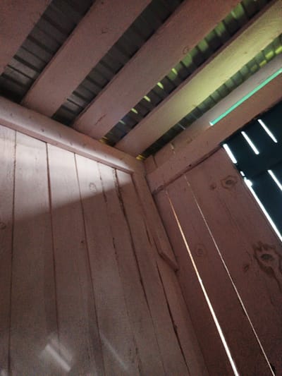 The image captures an interior view of a wooden structure, possibly a shed or barn, with a slatted wooden ceiling and planked walls. Sunlight is streaming through gaps in the structure, creating diagonal lines of light. The wood appears weathered and unfinished. There are no people or any identifiable objects present, other than the structural elements of the building itself. The location within the city of Islambaevo, Russia, is not discernible from the visual information in the image.