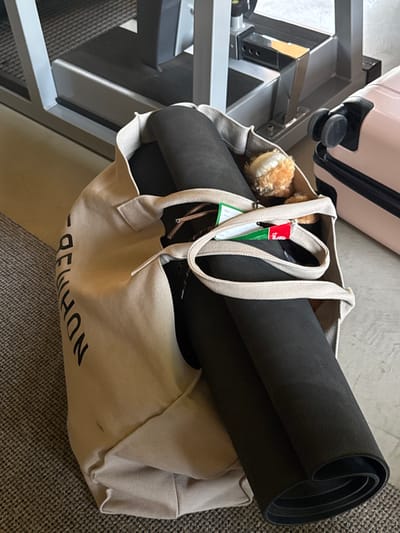 A beige tote bag lies on the floor, packed with a rolled-up black yoga mat, a stuffed animal, and a green tag attached to the strap. The bag is placed next to a gray, multi-tiered metal structure and a pink suitcase. The bag has "NDHMMON" printed on it, but the text is mirrored. The floor is covered in a carpet that appears to be gray.
