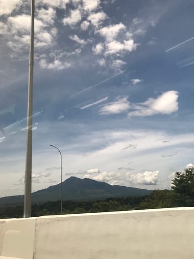 The image depicts a view through a window, reflecting the sky with white clouds. Outside, there's a mountain range covered in lush greenery and several light posts. A gray rail runs horizontally through the bottom portion of the image. Part of a white wall is on the right side. The location is likely Seulimeum, Indonesia, based on the implied geography.