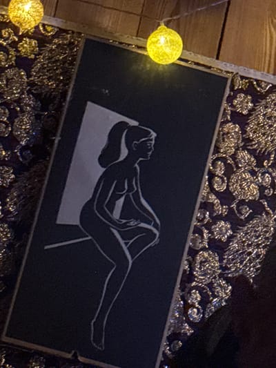 The image shows a close-up of a framed artwork, possibly a print or painting, featuring a silhouette of a woman in a crouching pose against a light-colored background. The artwork is set against a dark, textured fabric with intricate, shimmering patterns. Two illuminated spherical lights, resembling fairy lights, are visible in the upper portion of the image, casting a warm glow. The overall aesthetic suggests a decorative or artistic display. The location in Jāņupe, Latvia is not discernible from the provided image.