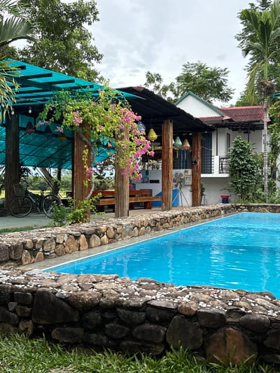 The image depicts an outdoor scene in Bố Trạch Commune, Vietnam, featuring a swimming pool with vibrant blue water. The pool is bordered by a stone wall, with a layer of smaller white stones on top. Beyond the pool, a covered outdoor seating area with a turquoise roof is visible, supported by wooden and corrugated metal posts. Pink bougainvillea vines are draped over the structure. In the background, a two-story building with a white facade and a reddish-brown roof is situated. Lush green trees and vegetation surround the area, with a clear sky overhead. A bicycle with a basket is parked to the left of the seating area. The overall impression is one of a tropical resort or guesthouse.