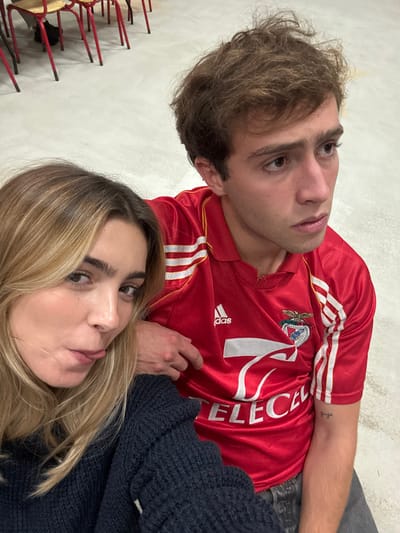 The image features two young adults in a close-up, indoor setting within Décines-Charpieu, France.
**Subjects:**
1. **Female (Left):** A young adult with blonde hair, parted on the side, wearing a dark blue, textured knit sweater. Her right arm rests on the male's left arm/shoulder. She looks directly at the camera with a playful expression, mouth slightly open, and tongue partially extended.
2. **Male (Right):** A young adult with short, disheveled brown hair, wearing a red short-sleeved Adidas football jersey. The jersey displays white stripes on the shoulders, an Adidas logo, the S.L. Benfica club crest, the sponsor "TELECEL," and the number "7" in white. The collar shows minor wear. A small tattoo "guga" is present on his inner left forearm. He looks off to his right (viewer's left) with a serious or pensive expression.
**Interaction:**
The female is leaning into the male, with her arm placed across his, indicating proximity.
**Environment:**
The background consists of a light-colored, plain floor. In the upper left corner, several chairs with red metal frames and light-colored seats/backs are visible, arranged in a casual manner. This utilitarian environment suggests an indoor public or institutional space.
**Location:**
This indoor scene is located in Décines-Charpieu, France, within a facility consistent with a community hall, school, or multi-purpose room.