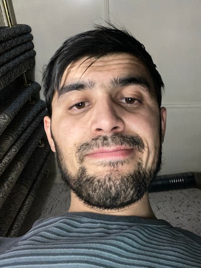 A person with dark hair and a beard looks directly at the camera. Their skin appears fair, and they have brown eyes. The person is wearing a gray striped shirt. Behind them, there is a curtain with a dark pattern and what looks like a ribbed metallic object. The image is rotated to the side.