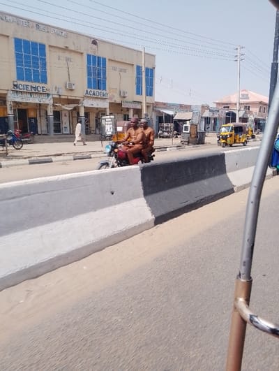 This image captures a daytime street scene in Chiranchi, Nigeria, characterized by urban infrastructure and activity.
In the immediate foreground, a concrete road divider, painted white with a black upper section, separates the paved asphalt roadway. A metallic pole, belonging to the vantage point, is visible on the far right edge.
Midground elements include two males, wearing matching brown attire and hats, riding a red motorcycle leftward on the road alongside the divider. Further down the street, two yellow auto-rickshaws are positioned on the right side of the road.
The background on the left is dominated by a two-story commercial building featuring a light-colored facade and multiple blue-tinted, gridded windows. Ground-level signage includes "SCIENCE" and "ACADEMY," with additional visible text such as "COMPUTER B.B.S.L STUDIO," "POS," and "AL GLOBAL VENTURES." Air conditioning units are mounted below the windows. A male pedestrian in a light-colored robe walks leftward on the sidewalk in front of this building, where a red motorcycle is parked.
To the right of the main building, the street continues with smaller commercial structures. Overhead power lines span across the upper portion of the frame against a clear, pale blue sky. Further in the background on the right, a building with a light-colored roof and an "L. HAMID ACADEMY" sign is visible. Utility poles support the overhead lines.