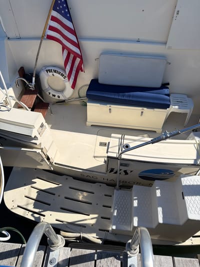 The image shows the stern of a white boat docked in the Village of Sag Harbor, United States of America. An American flag is flying from a pole on the boat. A white life preserver ring with "PREMIUMS TWO" written on it is hanging on the side of the boat. There are blue cushions on a bench seat. A white storage box is under the bench. A metal ladder is on the side of the boat. The boat has "MRS HARBOR" printed on its hull, with a logo of blue waves. There are white steps leading up from the water to the boat. The boat is made of fiberglass and the deck is a light color. The water appears to be dark. Wooden planks are visible in the foreground, likely part of the dock. The lighting suggests a sunny day with shadows cast on the deck.
