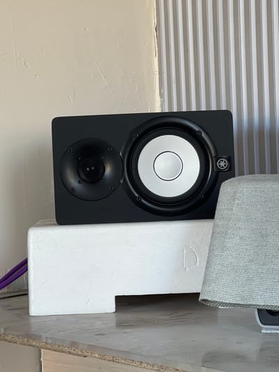 A black studio monitor speaker is positioned on a white styrofoam block. The speaker has two drivers: a smaller one on the left and a larger one on the right. The larger driver has a white cone and a black surround. The Yamaha logo is visible on the right side of the speaker. To the left of the speaker, purple cables are present. The speaker is situated on a countertop. Behind the speaker, a wall with vertical ribbed paneling is visible on the right, and a textured, off-white wall is on the left. A portion of a grey fabric object is visible to the right of the speaker. The image is taken indoors in a dimly lit environment. The location of Hazorasp, Uzbekistan is not depicted in the image.