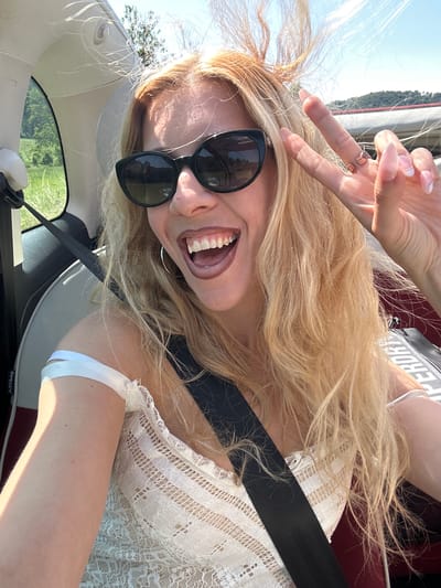 A blonde woman with sunglasses and a white lace top is sitting in the passenger seat of a car, making a peace sign with her right hand. Her hair is being blown by the wind, and she is smiling with her mouth open. The car appears to be moving on a road in Schoenfels, Luxembourg, with green trees and hills visible in the background. The sky is clear and blue.