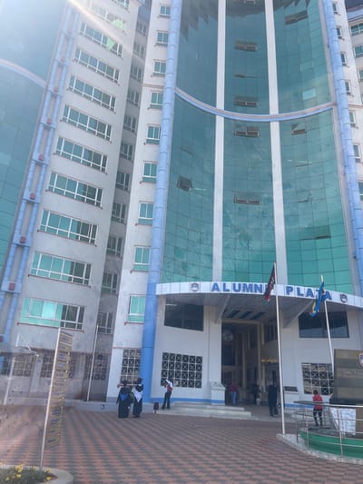 The image displays a tall, multi-story building identified by a sign over its main entrance as "ALUMNI PLAZA." While the provided context specifies a hospital ward in Kenya, the visual evidence indicates a university or institutional building.
The structure features a light-colored facade punctuated by numerous rectangular windows. Portions of the building, particularly the central section, are clad in reflective blue-green glass panels arranged in a grid pattern, with some sections exhibiting a curved architectural design. Vertical blue columns or decorative strips accent the building's exterior.
At ground level, a broad entrance with steps leads up to a recessed doorway, sheltered by a curved canopy that prominently displays the "ALUMNI PLAZA" lettering and associated emblem. Directly in front of the entrance stand two flagpoles; one flies a flag with red, black, and white stripes (consistent with the Kenyan flag), and the other displays a flag with blue and yellow colors.
The foreground consists of a paved area with a distinct checkerboard pattern of reddish-brown and darker gray or black tiles. To the left, a tall, vertical signpost with multiple directional arrows is visible. To the right of the entrance, a low monument or barrier with a green metal railing is present.
Approximately eight individuals are visible around the entrance area. Two individuals on the left are dressed in academic gowns and caps, suggesting participation in a graduation event. Other individuals are observed walking or standing near the entrance, including a person in a red top to the right of the steps. The scene is brightly illuminated by daylight, with a noticeable sun glare in the upper left portion of the image.
