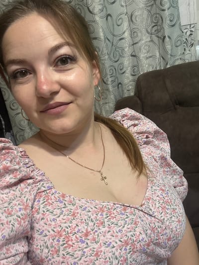 The image is a close-up, selfie-style photograph of a young woman with light brown hair and green eyes. She is wearing a pink floral print dress with puff sleeves and a gold necklace with a cross pendant. She is also wearing hoop earrings. The background consists of a patterned curtain and a dark brown upholstered chair. The scene appears to be indoors, possibly in a residential setting, in the city of Zubov, Russia.