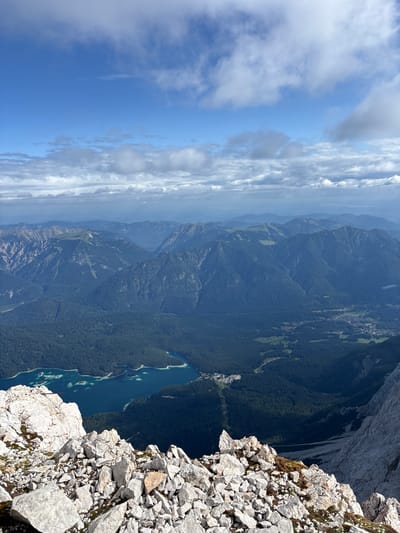 The image depicts a panoramic view of a mountainous landscape, with a bright blue sky dotted with white clouds. In the foreground, a rocky, grey terrain with sharp, irregular stones dominates the lower part of the frame. A vibrant turquoise lake is visible in the middle ground, surrounded by dark green, densely forested mountains. Further in the background, layers of progressively lighter blue mountains recede into the distance. A small town or village with scattered buildings and green fields can be seen nestled in the valley. The overall scene suggests a high-altitude vantage point, offering a vast and natural vista. The scene is characteristic of the Alps, and given the mention of Ehrwald, Austria, it is likely a view from a mountain peak in or near the Ehrwald region. No people or specific actions are discernible in the image.