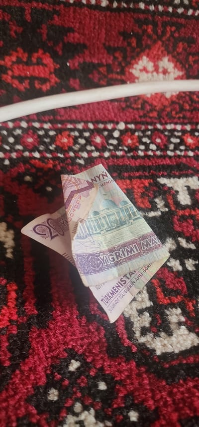 The image shows two Turkmenistani banknotes partially folded and lying on a red, black, and white patterned rug. One banknote, visible on the left, has the numeral "20" prominently displayed in purple. The other banknote, layered on top and to the right, features an illustration of a building with domes and arches, and text that reads "YGRiMi MAN" and "TÜRKiMENiSTAN." The banknotes are the primary subjects. The background is the textured surface of the rug. The scene is static and appears to be indoors. No people or specific locations within the city of Türkmenbaşy, Turkmenistan are depicted or implied.