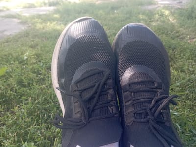 This is a close-up, eye-level shot of a pair of black athletic shoes, possibly running shoes, resting on a patch of green grass. The shoes appear worn, with visible creases on the toe box and a textured mesh material on the upper. Black laces are tied in a bow on each shoe, and the soles are a light color, perhaps white or cream. The background is softly focused, showing more grass and a hint of a lighter path or dirt area in the distance, suggesting an outdoor setting. The lighting is bright, indicating daylight, possibly with some dappled sunlight filtering through. There are no people visible in the frame, only the shoes and the natural environment.