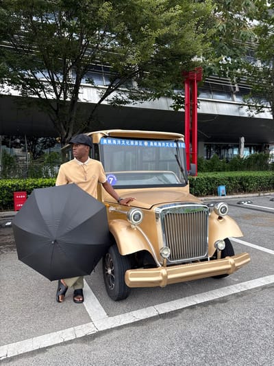 An adult male stands on an asphalt surface next to a gold-colored, vintage-style electric vehicle in the Olympic Sailing Marine Culture Tourism Zone, Shinan District, China. The man, with a dark complexion, is dressed in a tan short-sleeved polo shirt, khaki pants, black sandals, and a dark flat cap. He holds a closed black umbrella in his left hand, which rests on the ground. His right hand is placed on the gold-painted front fender of the vehicle.
The gold vehicle features a prominent chrome vertical-slat grille, circular headlights, and a gold-colored bumper. Its windshield displays the Chinese characters "奥帆海洋文化旅游区" (Olympic Sailing Marine Culture Tourism Zone) in blue text, repeated, and a white circular sign with the number "7". The asphalt surface beneath shows white parking space markings.
In the background, dense green foliage from trees partially obscures a multi-story modern building with reflective windows. A tall, red, rectangular structure composed of stacked horizontal elements stands behind the vehicle's right side. Green bushes line the edge of the paved area, and a red fire extinguisher cabinet is partially visible on a wall in the left mid-ground. The lighting is even, suggesting an overcast day.