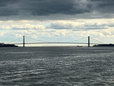 The image shows the Verrazano-Narrows Bridge spanning the Narrows strait in New York City. The view is likely from the vicinity of Bayonne, New Jersey. The scene features a large body of water with visible waves in the foreground. The bridge, a double-decked suspension bridge, stretches across the water connecting Staten Island and Brooklyn. The sky is overcast with dark, heavy clouds above lighter, more scattered clouds. There is a small vessel visible on the water near the bridge and a navigation buoy. Two landmasses frame the bridge on either side. The picture portrays a grey, slightly somber atmosphere.