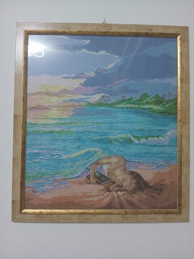 A framed cross-stitch depicting a sunset over the ocean with a person lying face down on the beach. The sky is filled with clouds in shades of blue, purple, yellow, and white. The ocean is a vibrant turquoise with white waves crashing on the shore. The beach is sandy brown. The person is lying with their head down, their arm bent, and their hair spread out on the sand. They appear to be holding a dark object in their hand. The artwork is hanging on a white wall. The frame is made of light-colored wood with a gold inner border. The image does not contain any information that would indicate its location in Monsummano Terme, Italy.