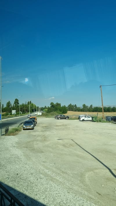 A wide-angle shot captures an outdoor parking area on a clear, sunny day in Larissa, Greece, likely near a road. The sky is a vibrant, cloudless blue. In the foreground, a gravel parking lot dominates the scene, with a shadow of a lamppost stretching across it. To the left, a paved road runs parallel to the parking lot, with a few cars parked along its edge. The cars include a dark sedan, a yellow minivan, and a silver hatchback. Further down the road, more vehicles and streetlights are visible, along with a verdant line of trees and bushes. In the middle ground, several cars are parked in the gravel lot, including a silver sedan, a white minivan, and a brown SUV. A green dumpster is situated to the right of the white minivan. In the background, a field of dry, golden grass stretches towards a line of trees, with mountains faintly visible in the distance. The scene appears to be a relatively open and somewhat rural area within or on the outskirts of Larissa.