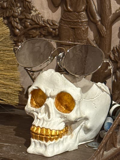 The image shows a close-up of a white skull adorned with gold accents in the eye sockets and along the jawline. Silver-rimmed sunglasses hang from the skull. The skull is positioned against a dark wooden background with carved figures of people. A bundle of straw is visible below the skull and glasses. The image was taken in Kladovitsy, Russia.
