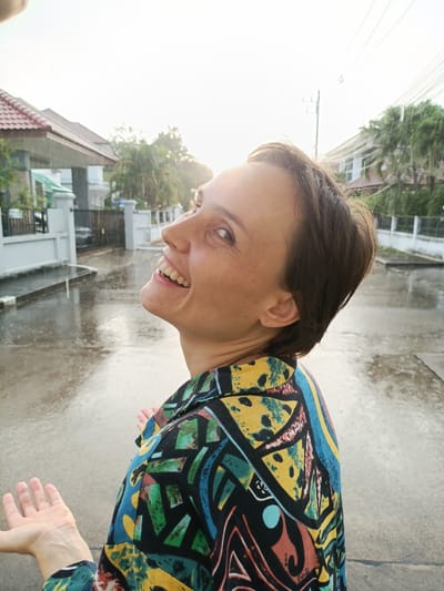 A woman is standing outside in the rain in Ban Patan, Thailand. She is looking up at the sky with her mouth open in a smile, her arms outstretched. She is wearing a colorful patterned shirt. The street is wet and reflective, and there are houses and trees in the background. The sun is shining through the rain, creating a bright, hazy effect.