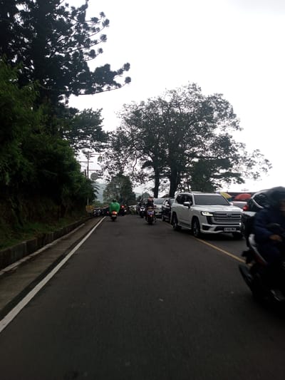 The image shows a congested road with cars and motorcycles in Mae Chan District, Thailand. The vehicles are moving slowly or stopped in traffic. On the left side of the road, there is a concrete curb separating the road from a grassy hillside with trees. A white line marks the edge of the road. Trees are visible on both sides of the road, and the sky is overcast. The traffic includes a white SUV with license plate "A 666 UKK".