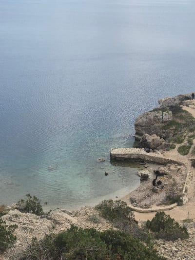 This image presents an elevated, downward-looking view of a rocky coastal bay within the Municipal Unit of Loutraki - Perachora, Greece. The upper two-thirds of the frame display a calm, blue-grey body of water with minimal surface ripples. Closer to the shore, the water appears clear, revealing a lighter blue-green hue over a sandy or gravelly seabed.
The lower third of the image encompasses a rugged, arid coastline composed of light-colored rocks and dirt paths. Sparse, low-lying green vegetation, primarily bushes, is scattered across the foreground and rocky slopes.
A stone pier, constructed from light-colored blocks, extends approximately 15-20 meters into the water from a small, sandy cove located towards the center-right of the lower frame. Adjacent to the pier, a curved beach area with light-colored sand and gravel lies at the base of the rocky embankment. Stone retaining walls or pathways are integrated along the curve of the beach and ascend the adjacent slopes.
One human figure, dressed in dark clothing, is visible standing on a dirt path atop a rocky cliff in the upper right section of the landmass, overlooking the water. No specific actions are discernible from this distance.
On the sandy area near the base of the cliffs, a yellow object, possibly a container, is present. Nearby, irregular dark patches are observed on the ground. A dark, barrel-shaped object rests on the beach immediately beside the pier. The scene is illuminated by natural daylight.