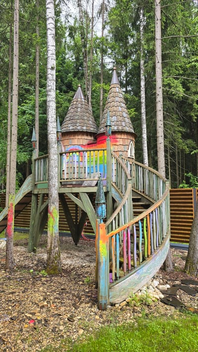This is a vertically oriented, full-shot, eye-level, outdoor photograph of a colorful, elaborate treehouse-like structure, resembling a castle. The structure is predominantly made of wood, with two prominent conical roofs tiled with what appears to be small, dark shingles. The sides of the structure are decorated with colorful wooden planks arranged in a rainbow pattern, and some parts feature intricate carvings. A wooden staircase with rainbow-colored steps leads up to a curved walkway. The treehouse is situated in a wooded area with tall, thin trees and lush green foliage in the background. The ground around the structure is covered with small stones and pebbles, with patches of green grass. The overall impression is whimsical and playful, suggesting it is a child's play area. The lighting suggests it is daytime, with diffused light indicating an overcast or possibly early morning/late afternoon setting. There is no visible text in the image. The location context is Kamenka, Russia.