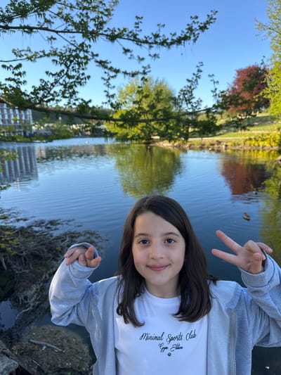 A cheerful young female, likely a child or pre-teen, is captured in a sunny outdoor portrait, smiling towards the camera. She has long dark hair and is wearing a white t-shirt with the text "Minimal Sports Club Gym Edition" and a small dumbbell icon, layered under a light grey hoodie. Her left hand is raised, forming a peace sign, while her right hand shows a similar gesture.
Behind her is a calm body of water, appearing to be a pond or river, which reflects the clear blue sky above. Scattered ducks are visible on the water's surface. The edges of the water are lined with lush green trees, some with verdant foliage and others displaying reddish-brown leaves. Reflections of modern buildings can also be seen in the water, suggesting an urban park or waterfront setting. The sky is bright and clear, indicating a pleasant, sunny day. The overall scene depicts a serene natural environment within a city. This location is in Ulm, Germany.