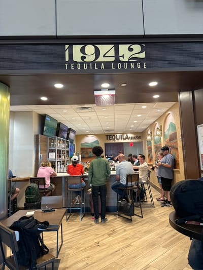 The image depicts the 1942 Tequila Lounge inside Tampa International Airport in Tampa, Florida, United States. The lounge is visible with its bar area populated by several individuals. Some patrons are seated at the bar, while others are standing or seated at tables nearby. The lounge is decorated with artwork and features a display of various tequila bottles behind the bar. Luggage is visible near the seating area, indicating that patrons are likely travelers. Televisions are positioned above the bar, presumably showing entertainment or news. The lounge's entrance is marked with a prominent sign bearing the "1942 Tequila Lounge" name. The environment suggests a busy airport atmosphere.