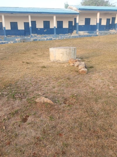 This image depicts an outdoor scene in Okeni, Nigeria, featuring a desolate field in the foreground and a public building in the background. The ground is covered by dry, patchy brown and green grass. A cylindrical concrete wellhead, approximately waist-height, is situated in the midground, with faint, weathered text visible on its side, including "PROJECT B.E.B" and "KOGI." A line of approximately six irregularly shaped, light brown rocks extends from the right side of the wellhead towards the foreground. Another larger, light brown rock is present in the lower-left foreground.
In the background, a single-story building with a light blue corrugated metal roof and white walls is visible. A dark blue horizontal band of paint runs along the lower portion of the building's facade, showing significant peeling and wear. The building features a series of dark blue rectangular windows and doors, along with white support columns creating a covered walkway or veranda. No individuals or active subjects are present in the frame. The overall environment suggests a dry, possibly arid, climate and an institutional setting, likely a school or community facility.