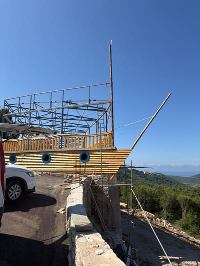 In Svinjišta, Montenegro, a wooden structure designed to resemble a ship is under construction. The ship-like building, adorned with blue circular windows, is situated on a hillside overlooking a body of water and lush green landscape. The structure's wooden plank exterior and railing are visible, along with a metal framework for the upper levels. A tall wooden mast extends upwards, supporting a thin white flag. Scaffolding surrounds parts of the building, indicating ongoing construction. In the foreground, a white car is parked on an asphalt surface, and a low stone wall with some debris is present. The sky is clear and blue.
