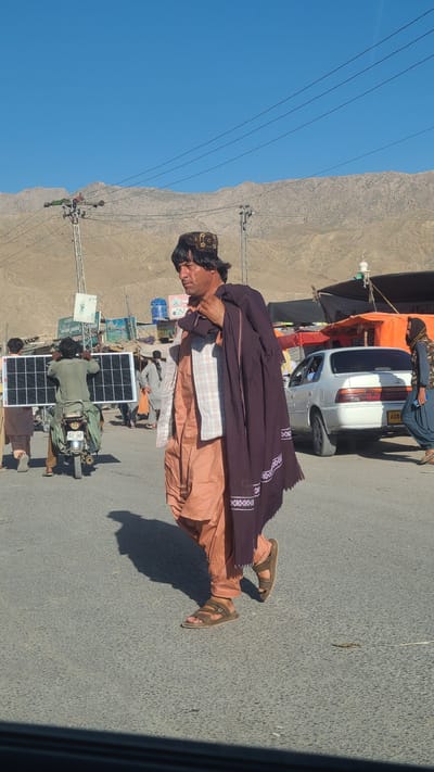 The image, shot in Bostan, Pakistan, shows a roadside scene on a bright day with a clear blue sky. A man in traditional Pakistani attire - a head covering, layered clothing, and sandals - is seen lying on the asphalt road. A dark shawl with embroidered patterns partially covers him. Behind him, a motorcycle equipped with two solar panels is visible, with additional figures nearby. A car is parked to the side under a makeshift awning. Power lines stretch across the top left corner of the frame, while a mountainous landscape serves as the backdrop.