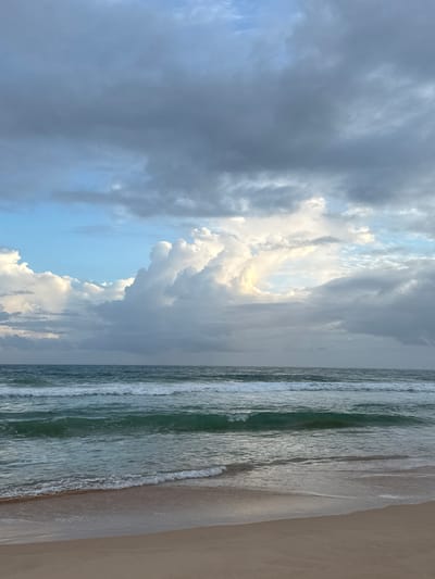 This is a vertical, eye-level, full shot of a beach scene in Koggala, Sri Lanka. The image is dominated by a dramatic sky filled with dark, moody clouds on the left, transitioning to bright, illuminated clouds with a hint of sunlight breaking through on the right. The ocean occupies the middle section, with gentle waves rolling towards a sandy beach on the far right. The water has a greenish hue and displays subtle patterns of foam as the waves crest and break. The sand appears damp and smooth from the receding tide. The time of day appears to be late afternoon or early evening, given the lighting and the dramatic cloud formations, which might suggest an approaching storm or the aftermath of one. The overall mood is serene yet powerful, capturing the natural beauty of the coastal environment. There is no visible text in the image.