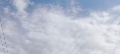 The image shows a cloudy sky with a few thin wires visible in the lower left corner. There are no people or discernible objects in the scene other than the wires and the sky. The clouds are predominantly white and grey, with patches of blue sky visible. The overall impression is that of an outdoor scene, likely taken during the daytime. There is no information in the image to suggest the location is Margilan City, Uzbekistan.