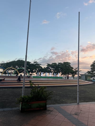 The image depicts a waterfront park in Bantaeng, Indonesia, during what appears to be late afternoon or early evening, given the soft lighting and colorful sky. In the foreground, a rectangular planter with lush green foliage sits on a tiled surface. Two tall, light-colored poles, likely flagpoles, are prominent in the scene, one on either side of the frame, with a section of their grey, textured surface visible.
The middle ground features a multi-tiered circular platform, illuminated by warm lights along its edges. Several people are present on this platform. Some are seated, while others are standing or walking. Behind the platform, large trees with dense green canopies dominate the landscape, providing shade. Across the water, which is visible beyond the trees, is a subtle hint of land or structures.
A notable feature of the park is a large, illuminated sign spelling out "SERUNI" in white, three-dimensional letters, positioned on the circular platform. To the left of the sign, a colorful arched structure, with yellow, green, and blue segments, adds a playful element to the environment. Further to the right, a small building with a light green roof and beige walls is visible. The sky above is a gradient of soft blue, with streaks of pink and white clouds, indicative of a sunset. The overall impression is a tranquil public space, likely a popular spot for recreation and relaxation in Bantaeng.