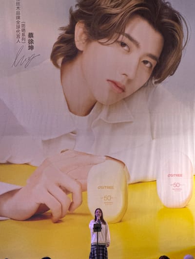 The scene depicts an indoor event in Changping District, China, centered around a large promotional backdrop. The backdrop features a prominent image of male celebrity Cai Xukun (蔡徐坤) from the chest up, with light brown and blond hair, wearing a white collared shirt, looking directly forward. Chinese characters on the upper left identify him as the global spokesperson for a brand's sunscreen series. His hands are visible at the bottom of his image, near two product containers. These products, labeled "OSITREE" with "SPF 50 PA++", are light yellow ("REPAIRING SUNSCREEN") and light orange/pink ("WHITENING SUNSCREEN"). The backdrop's lower section is bright yellow, transitioning to a light pink/white gradient at the top. In the foreground, positioned centrally against the backdrop, a young female stands at a microphone on a stand. She wears a white long-sleeved top and a dark plaid skirt, and her mouth is open, indicating she is speaking or singing. The tops of several heads are visible at the very bottom edge of the frame, suggesting an audience.