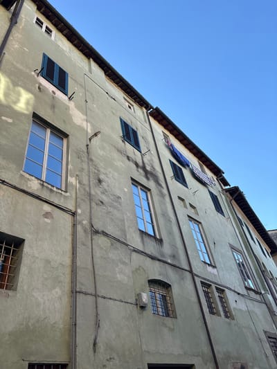 The image displays the weathered facade of a multi-story building in Lucca, Italy, viewed from a low-angle perspective against a clear blue sky. The building's exterior is surfaced with pale olive-green stucco, exhibiting areas of paint deterioration, dark vertical runoff stains, and lighter patches where paint has chipped.

Multiple windows are present across the facade. Several windows are equipped with dark teal louvered shutters, some of which are closed while others are partially open. Additional windows feature clear panes, reflecting the bright blue sky. On the lower levels, smaller rectangular windows are secured by dark metal grilles. An external white rectangular fixture, possibly a light or sensor, is mounted on the wall above one of these gridded windows.

Dark utility conduits and electrical wiring are affixed to the facade, running both vertically and horizontally. Several metal brackets protrude from the wall at various heights. Towards the upper right section of the building, blue and white-and-dark checkered fabrics are visibly suspended from an open window, indicating laundry drying. The building's dark-tiled roofline and gutter system are visible along the top edge of the structure. No individuals are depicted. The upper right portion of the frame is dominated by a clear, bright blue sky, with sunlight illuminating sections of the building's upper left facade.