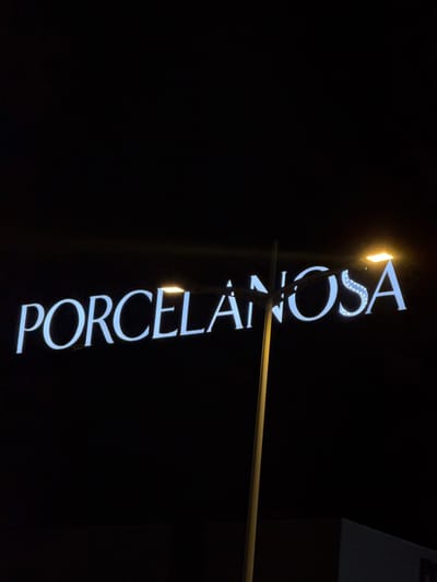 An outdoor, nighttime photograph depicts an illuminated "PORCELANOSA" sign in Polígon Industrial De Son Castello, Spain. The sign features white capital letters against a black background. A vertical, light-colored street light pole obstructs a portion of the letters "LAI" in the sign. Two active street lamps with bright, rectangular, yellow-orange light sources are visible: one above the "E" and "L" of the sign, and the other above the "O" and "S". A dark, angular building structure is discernible in the lower right corner of the frame. The sky is entirely black. No individuals or direct actions are visible.